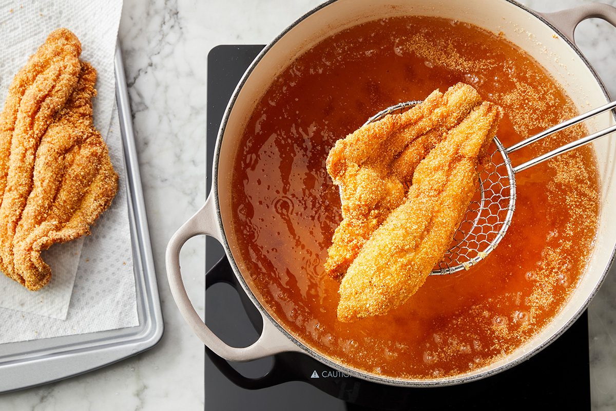 A breaded fish fillet is being lifted from a pot of hot oil with a metal strainer, while another cooked fillet drains on a paper towel nearby.