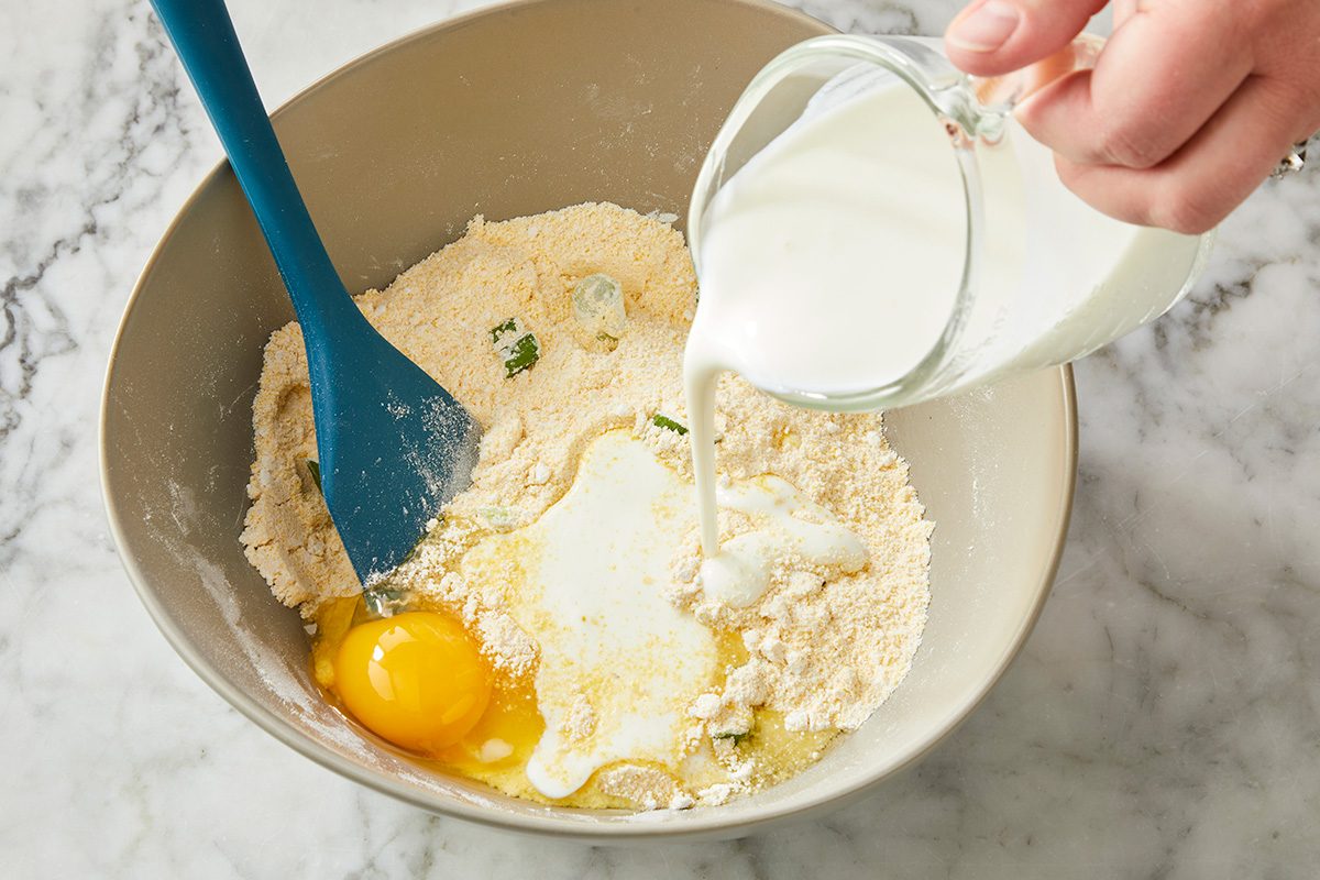 A hand pours milk from a glass measuring cup into a bowl with flour, an egg, and a blue spatula on a marble countertop.