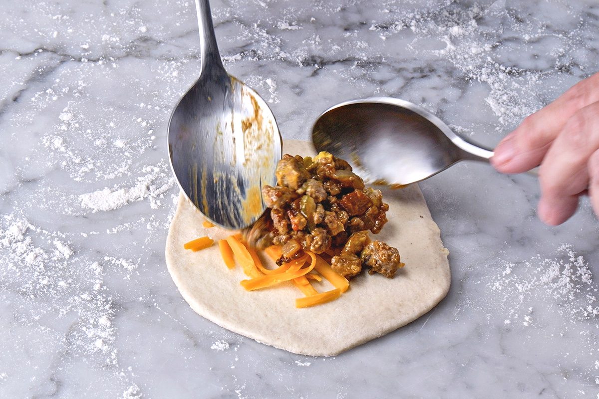 Two spoons are placing a mixture of cooked meat and orange shredded cheese onto a round piece of dough on a floured marble surface, preparing to fold and seal the dough.