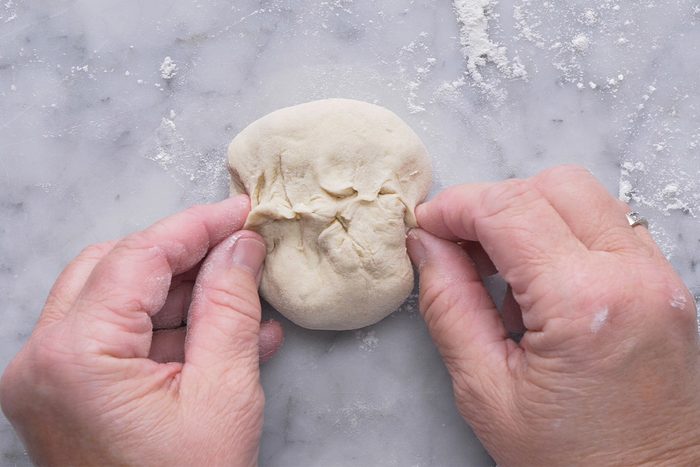 Two hands are shaping a small ball of dough on a lightly floured surface, pinching the dough together at the top.