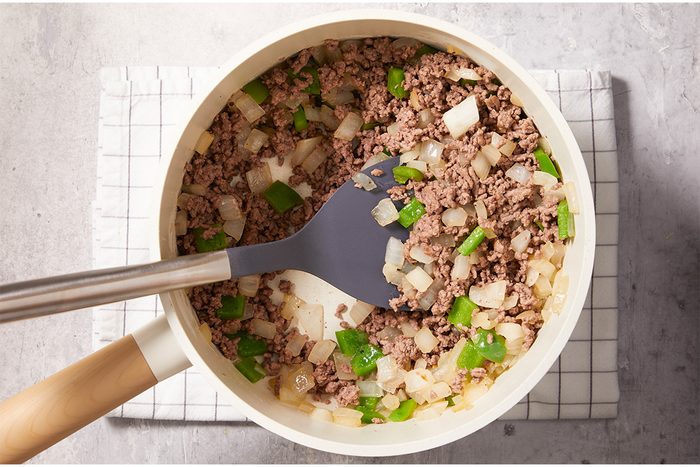 A pan filled with browned ground beef, chopped onions, and green bell peppers being stirred with a gray spatula, sitting on a white checkered cloth on a gray surface.