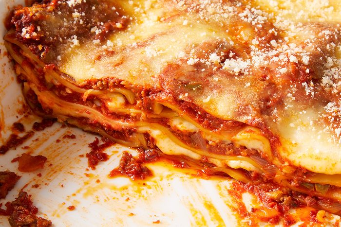 A close-up of a partially sliced lasagna in a baking dish, showing layers of pasta, meat sauce, and melted cheese with some crumbs and sauce around the edges.