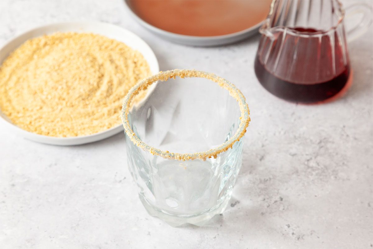 A clear glass with a crumb-coated rim sits on a light surface. Nearby are a plate of crumbs, a glass jug with red liquid, and a plate with a brownish liquid.