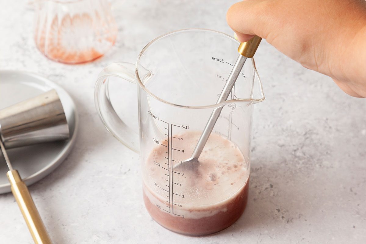 A hand stirs a pinkish liquid in a clear measuring cup with a gold-handled spoon, on a light countertop with a metal jigger and glass in the background.