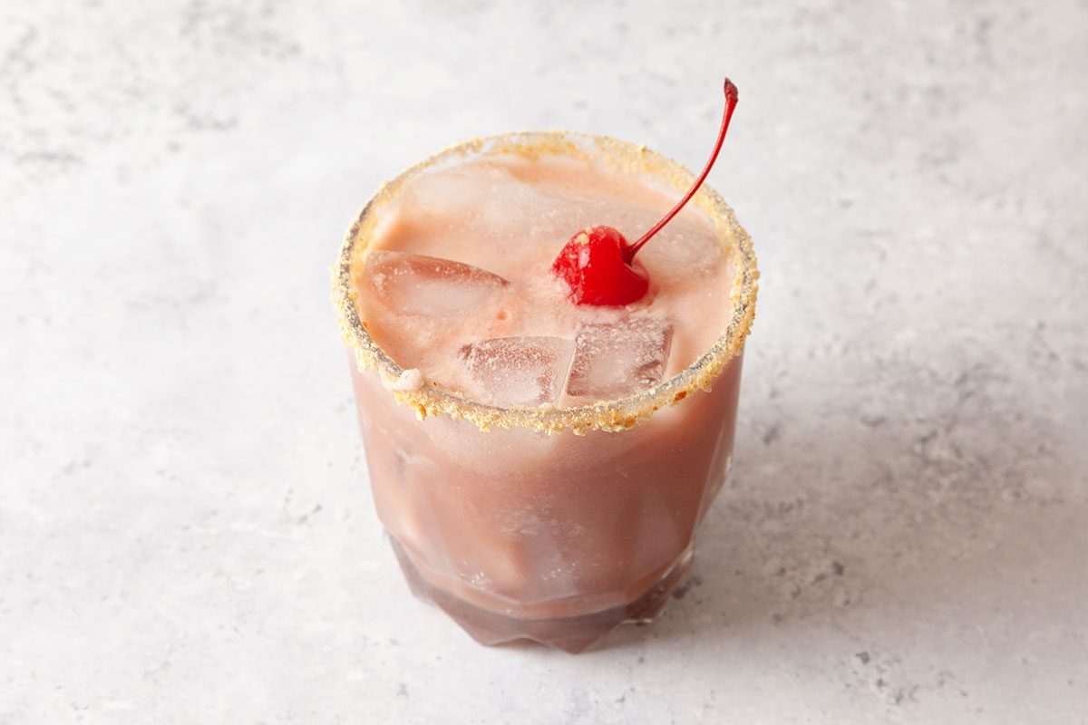 A glass of iced pink cocktail with a sugar-rimmed edge, garnished with a red maraschino cherry on top, sits on a light, textured surface.