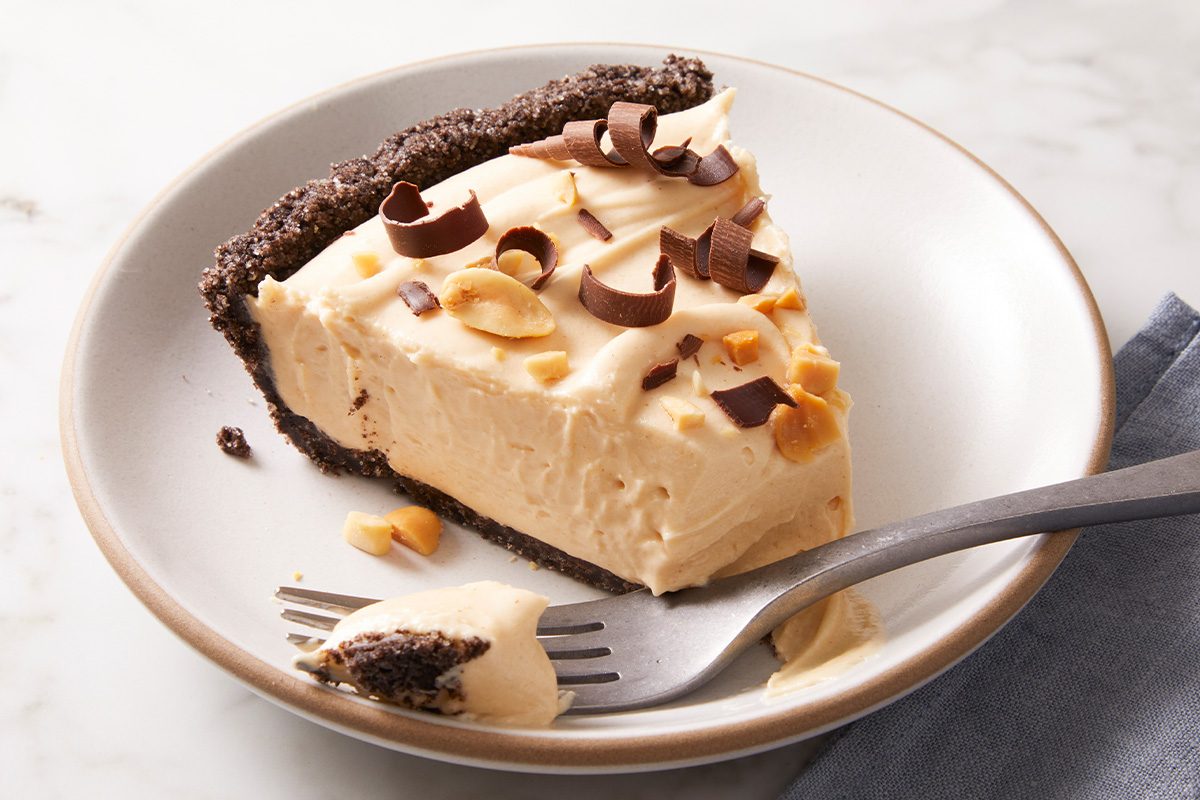 A slice of creamy pie with a chocolate crust, topped with chocolate curls and chopped nuts, sits on a white plate with a fork resting beside it.