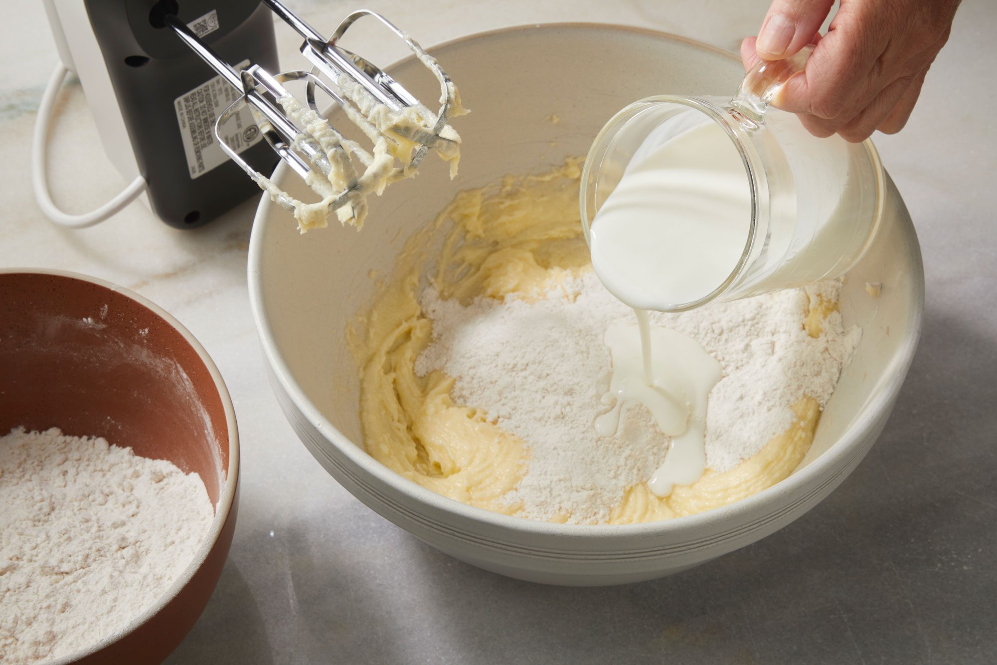3/4th shot of a hand pours milk from a glass measuring cup into a mixing bowl with flour and batter, while an electric mixer with beaters rests nearby, Another bowl with flour sits to the side on the countertop