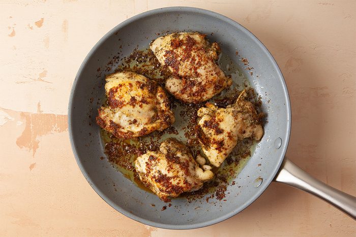 Four pieces of seasoned, browned chicken are cooking in a gray frying pan with some oil, set on a light-colored surface.