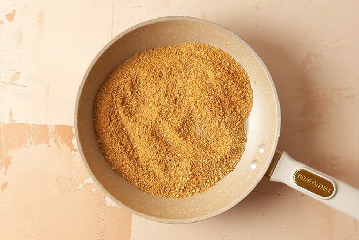 A white frying pan filled with toasted breadcrumbs sits on a light beige surface.