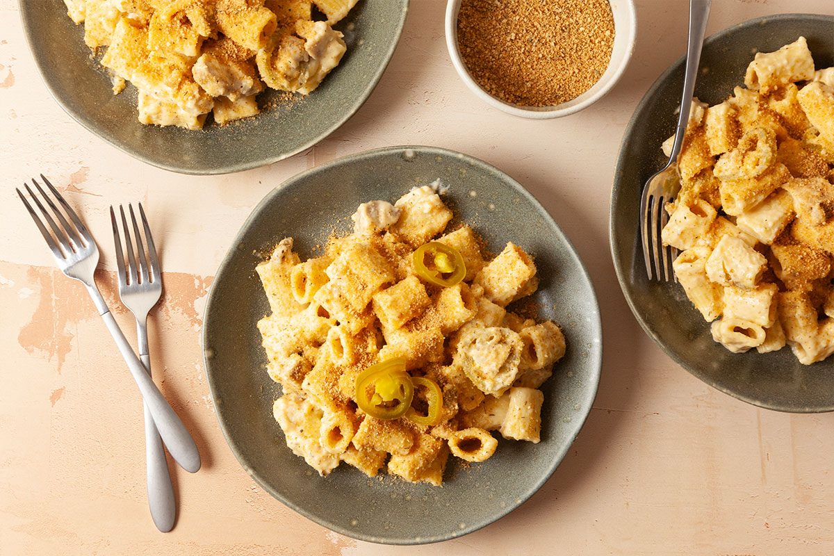 Three plates of pasta with creamy sauce, topped with breadcrumbs and sliced jalapeños, are arranged on a light table. A bowl of extra breadcrumbs and three forks are also visible.