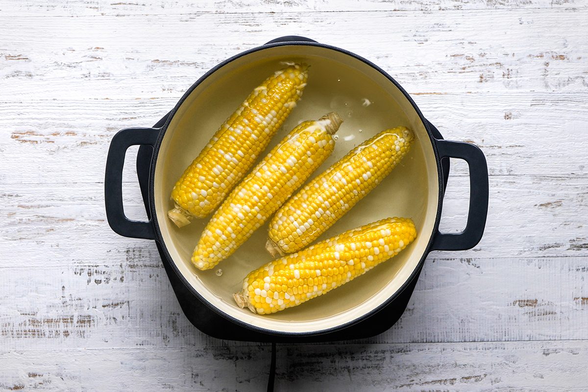 Four ears of corn are boiling in a pot of water on a stovetop, viewed from above. The background is a light-colored, rustic wooden surface.