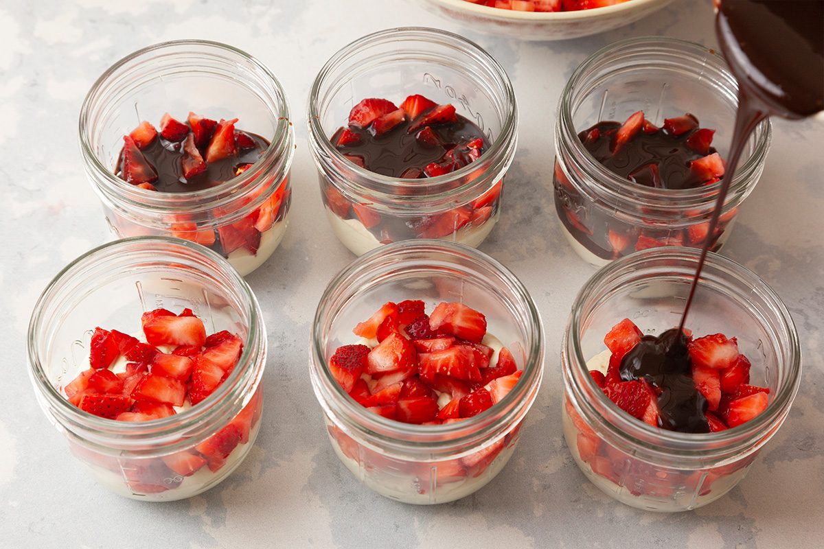 Six small glass jars filled with layers of cream and chopped strawberries, with chocolate sauce being poured onto the fruit in one of the jars.