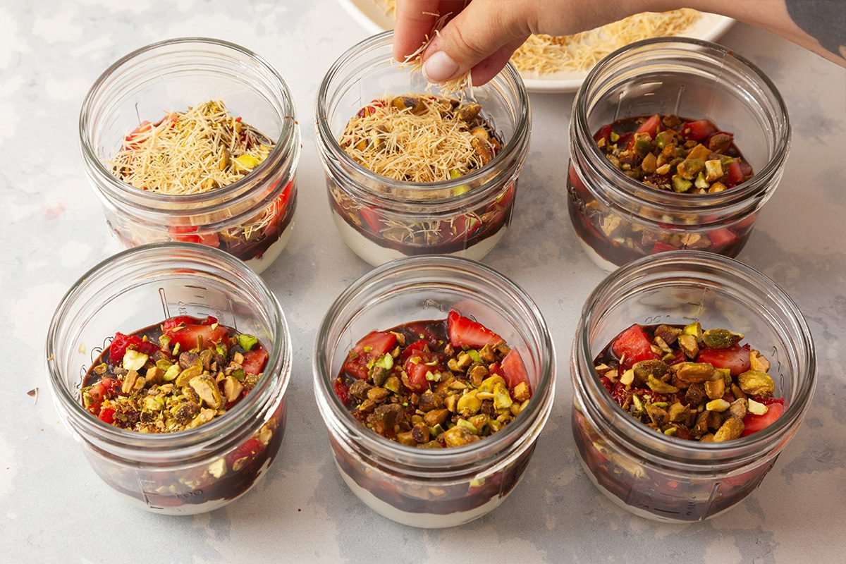 Six glass jars filled with layered desserts, topped with chopped nuts, strawberries, and shredded coconut. A hand is sprinkling coconut on one of the jars. The jars are arranged on a light-colored surface.