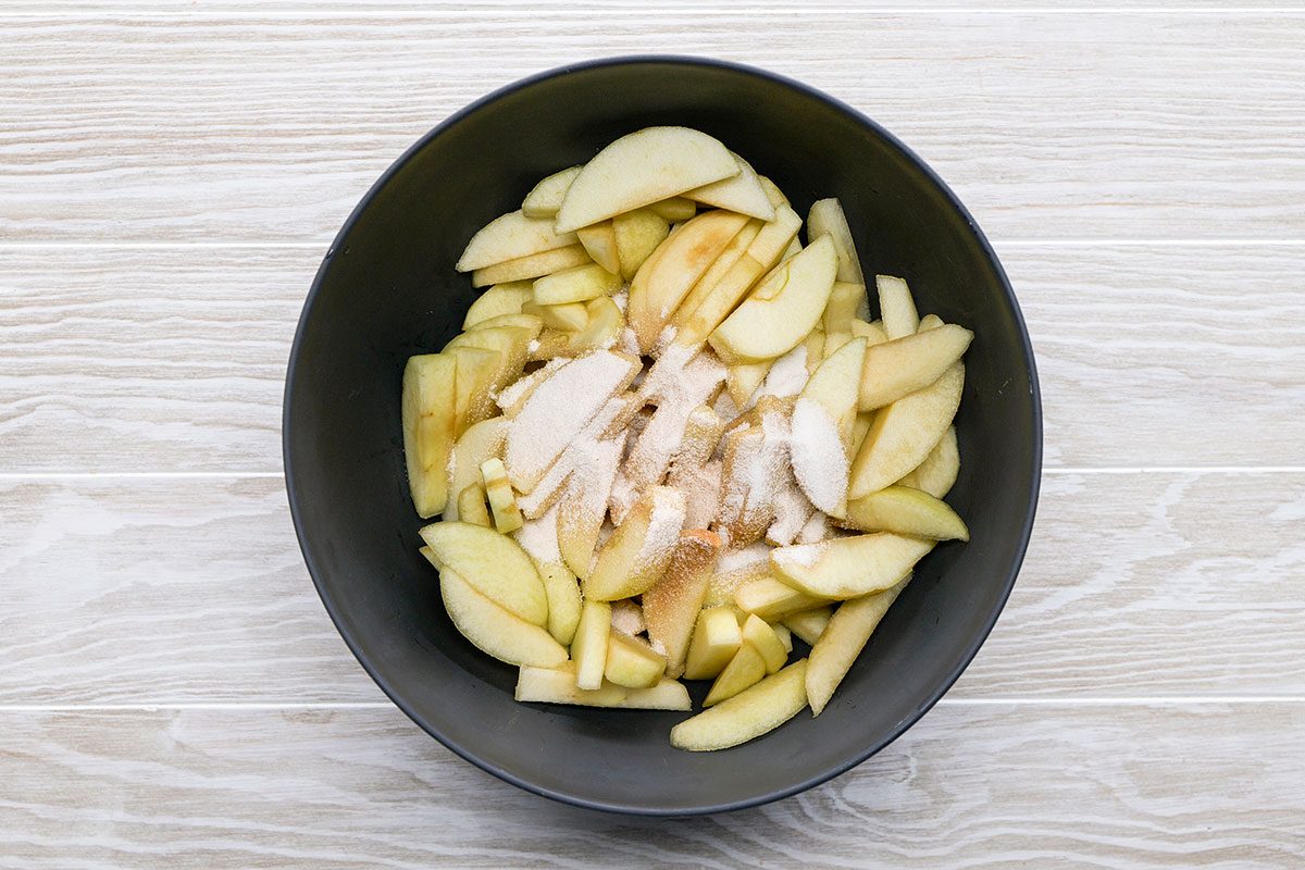 A black bowl filled with sliced apples and sprinkled with sugar and spices, placed on a light wooden surface.