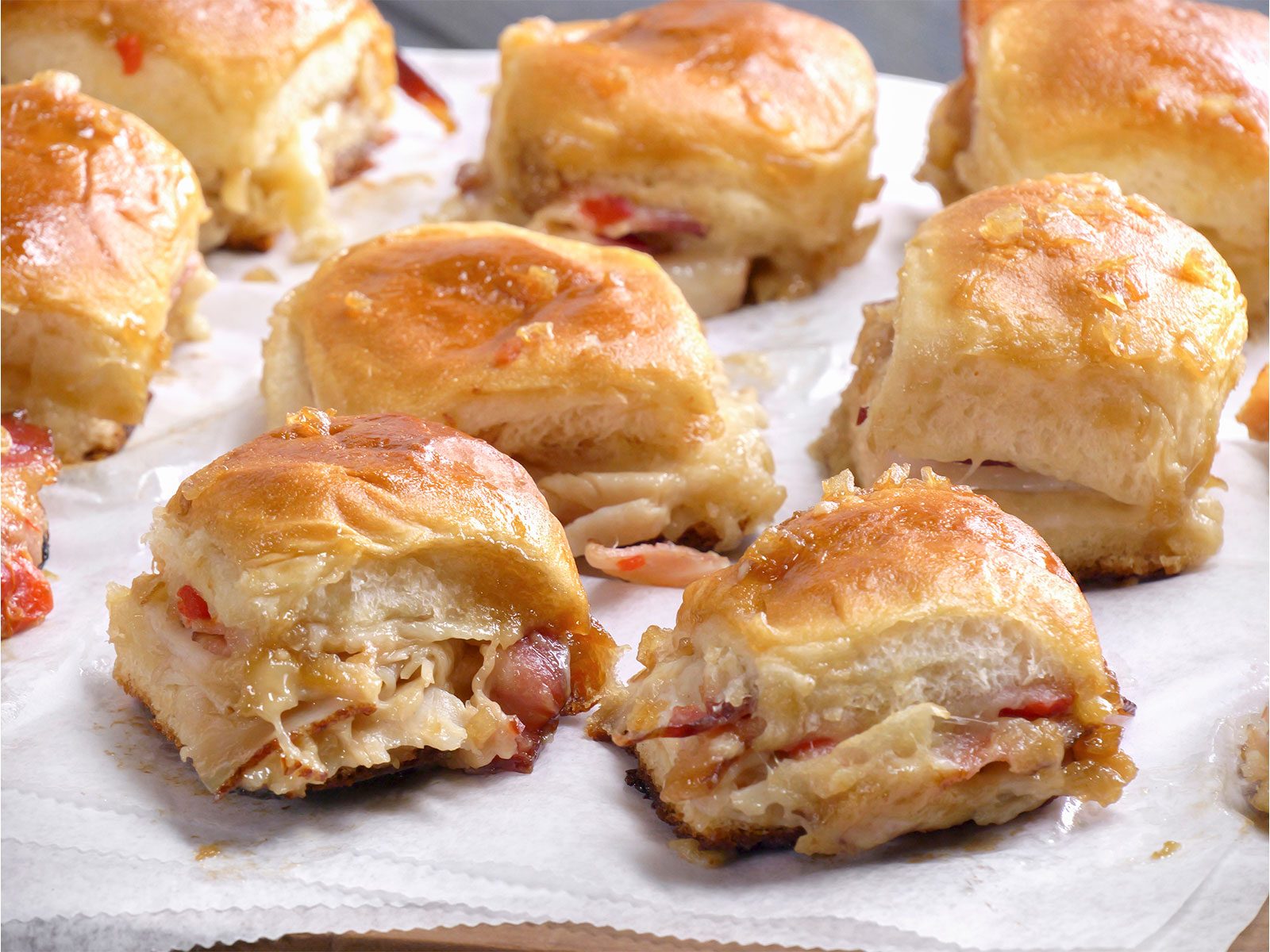 Close-up of several baked slider sandwiches with golden brown tops, filled with sliced deli meat and cheese, arranged on parchment paper