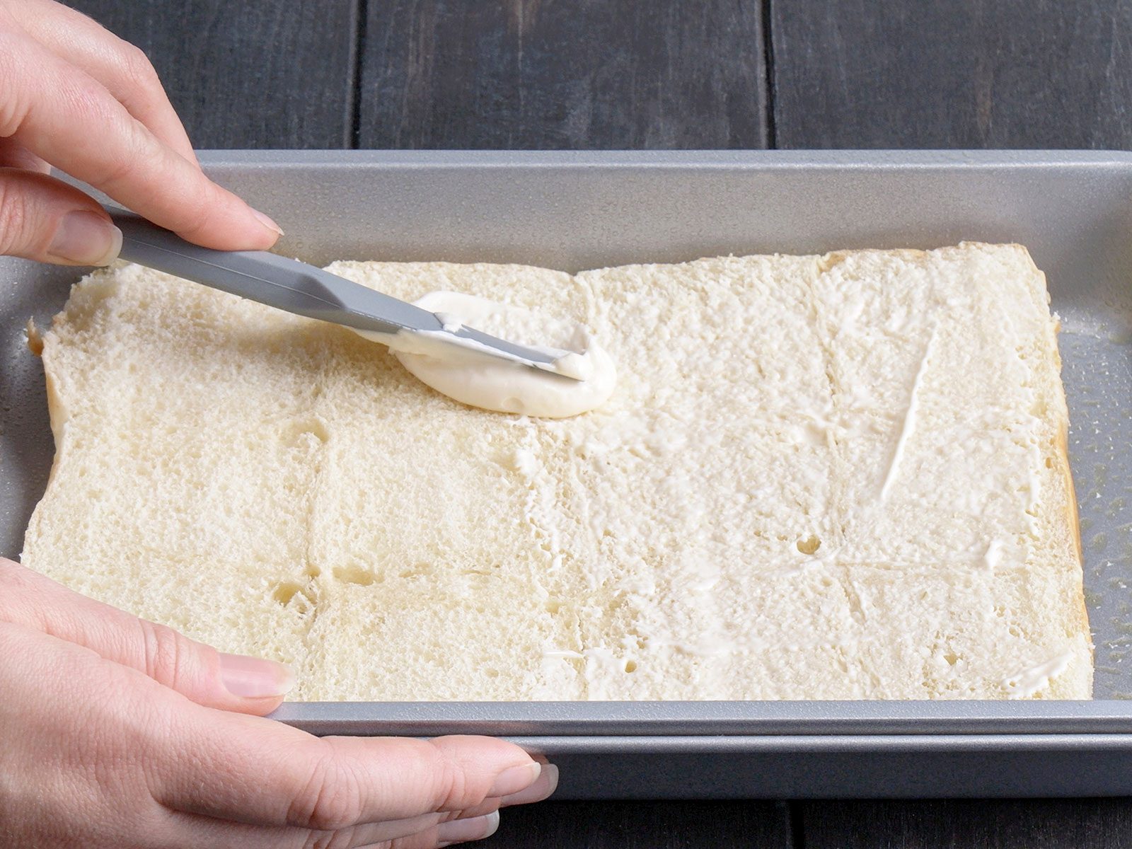 A person spreads a mayonnaise onto a layer of sliced bread in a rectangular metal baking pan using a spatula. The bread fills the bottom of the pan, and the surface is partially covered