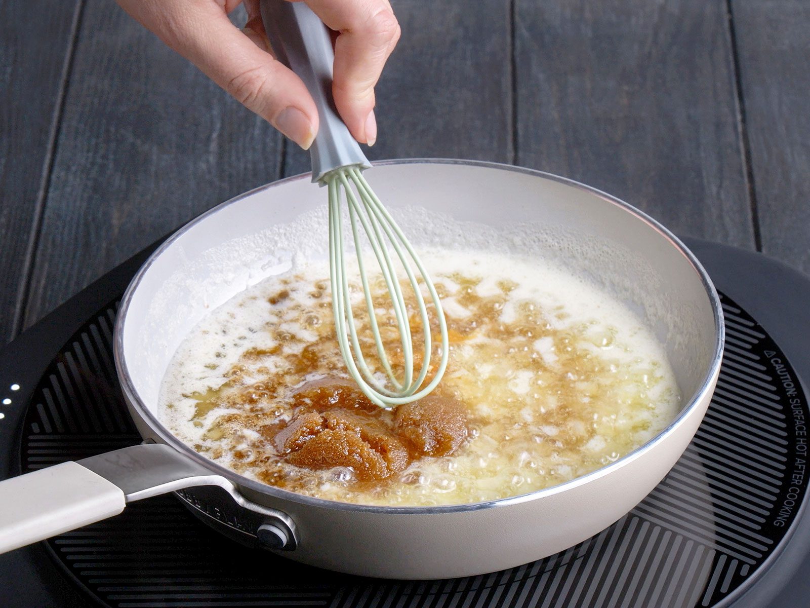 A hand uses a whisk to stir brown sugar into melted butter in a white saucepan on a stovetop, preparing a sauce