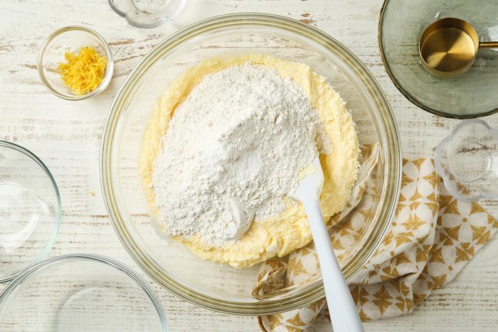 A mixing bowl with flour and creamed butter mixture being combined with a white spatula. Nearby are a small bowl of lemon zest, a glass with liquid, and an empty patterned kitchen towel.