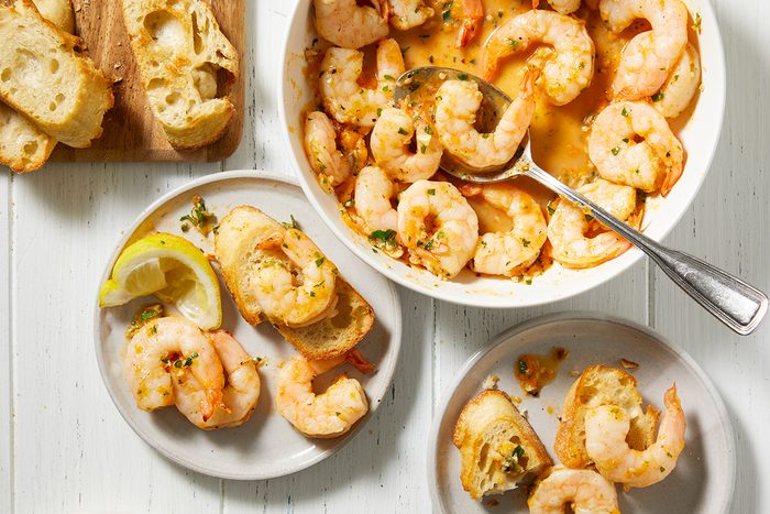 A white table with plates and a bowl of cooked shrimp in a garlic butter sauce. Slices of toasted bread are served alongside, some topped with shrimp. A spoon rests in the bowl. A lemon wedge is on one plate.