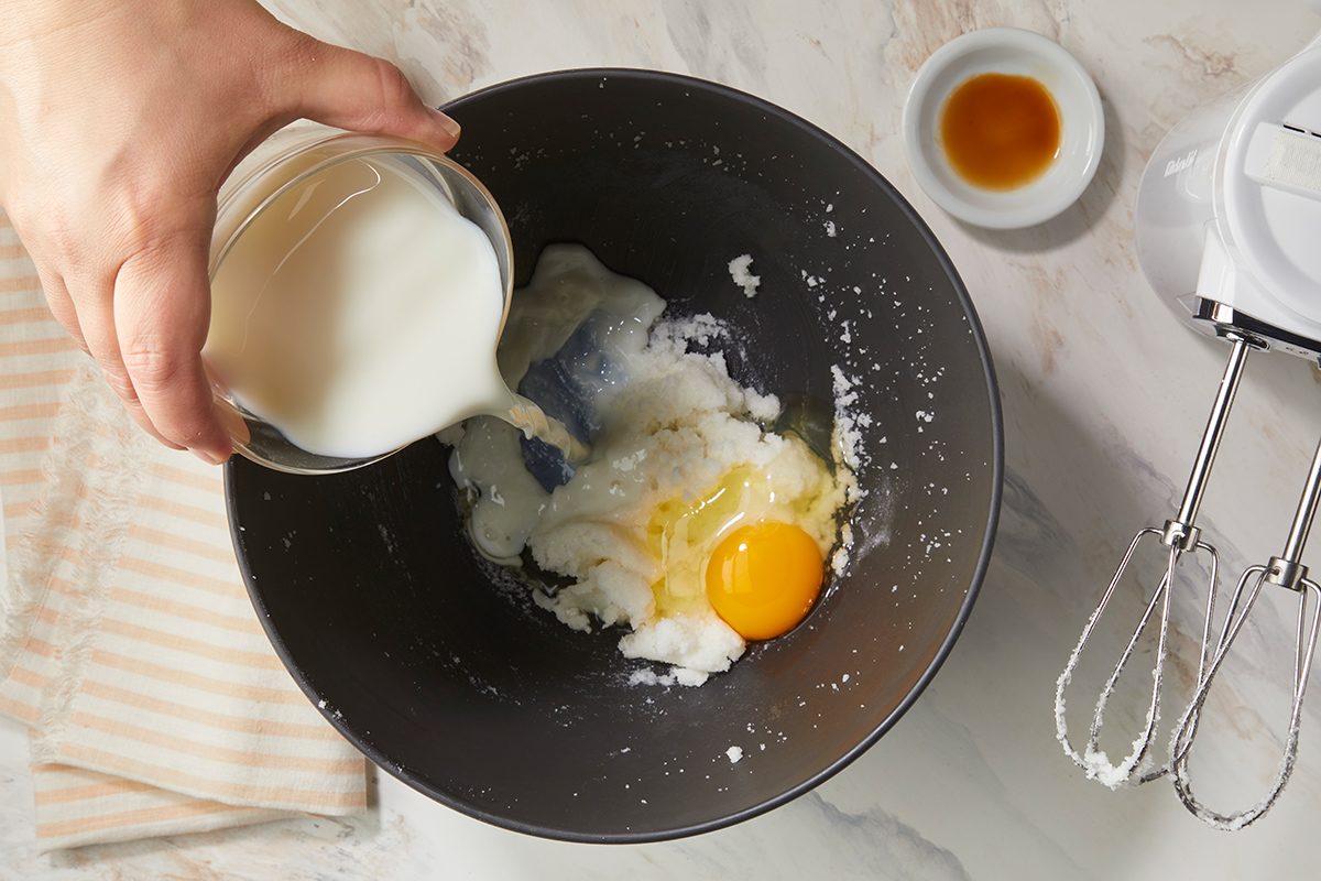 A hand pours milk into a mixing bowl containing sugar, an egg, and butter. Nearby are a striped towel, a small bowl of vanilla, and a hand mixer on a marble countertop.