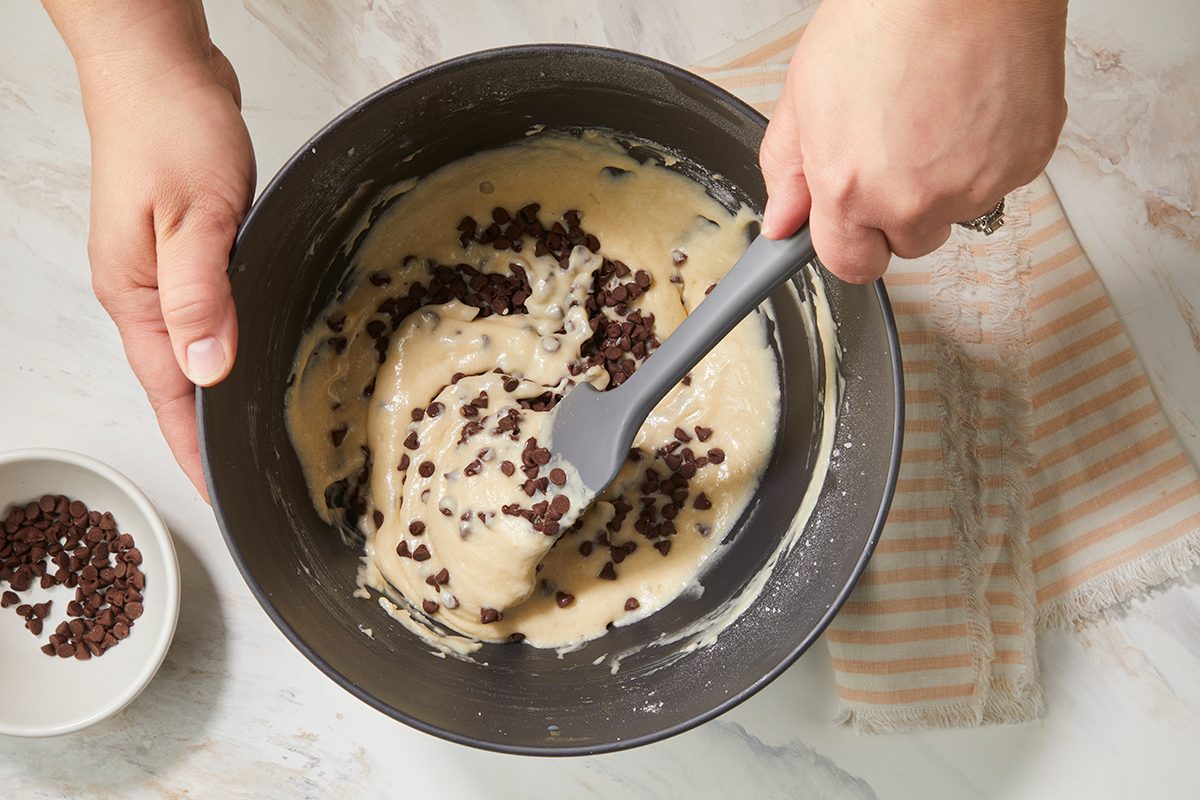 A person stirs chocolate chips into a thick batter with a spatula in a mixing bowl. A small bowl of more chocolate chips sits nearby on a marble countertop next to a striped cloth.