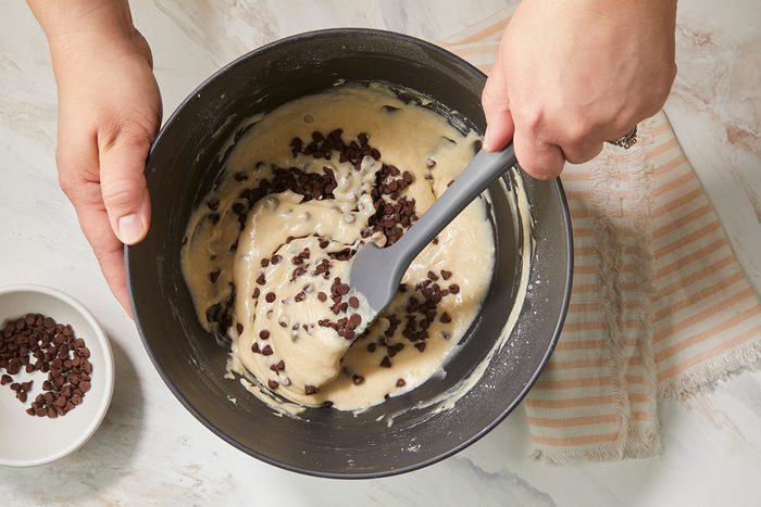 A person stirs chocolate chips into a thick batter with a spatula in a mixing bowl. A small bowl of more chocolate chips sits nearby on a marble countertop next to a striped cloth.