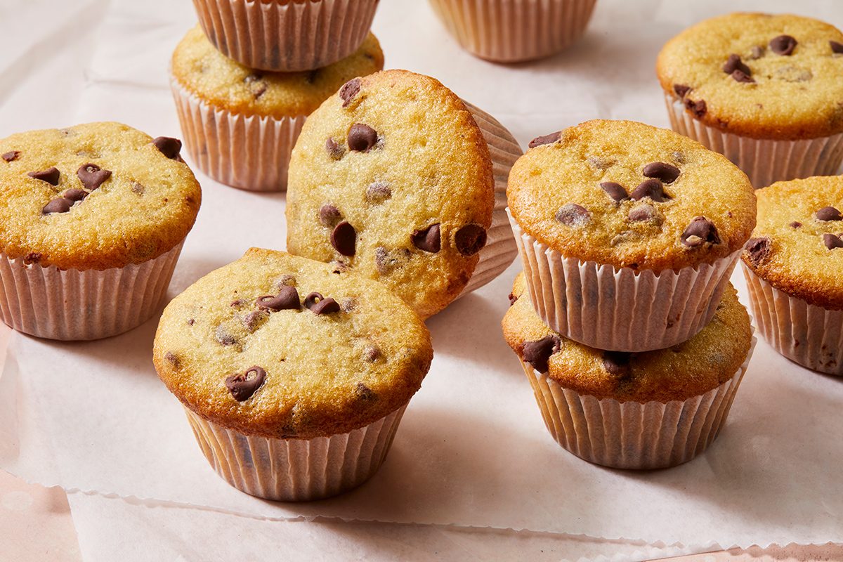 A group of chocolate chip muffins in paper liners are arranged on a white surface, with some muffins stacked on top of each other.