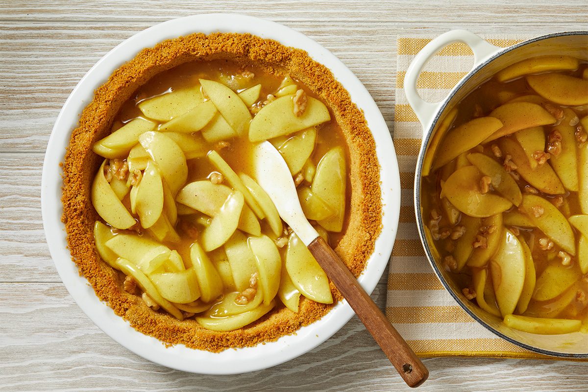 A pie dish with a crumb crust is being filled with sliced apples and walnuts in syrup. A spatula rests in the dish, and a pot with more apple filling sits nearby on a striped cloth.