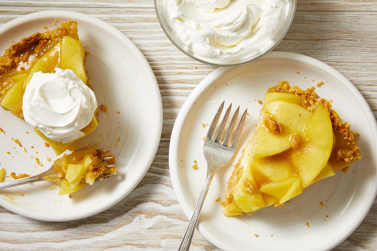 Two plates with slices of apple pie; one slice is topped with whipped cream and has a bite taken out with a fork. A bowl of whipped cream sits above the plates on a light wooden table.