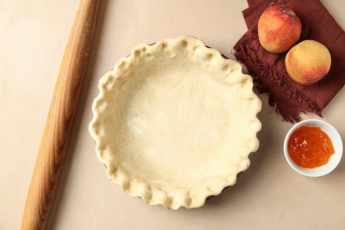 Unbaked pie crust in a pie dish next to a wooden rolling pin, two whole peaches on a brown napkin, and a small bowl of peach jam on a beige surface.
