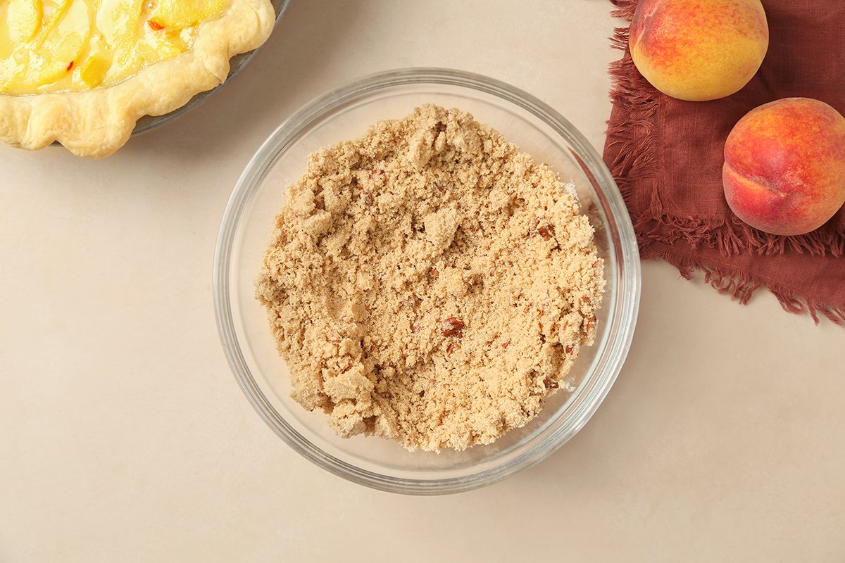 A glass bowl filled with brown crumb topping sits on a light surface near two peaches, a maroon cloth, and part of a baked pie.