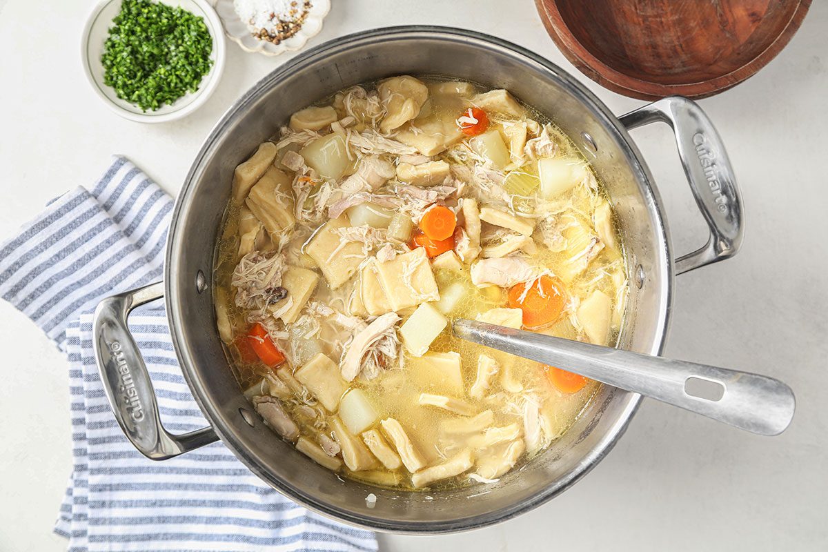 A pot of chicken and dumpling soup with visible chunks of chicken, potatoes, carrots, and dumplings. A spoon is inside the pot, with chopped herbs and a striped towel nearby on the countertop.