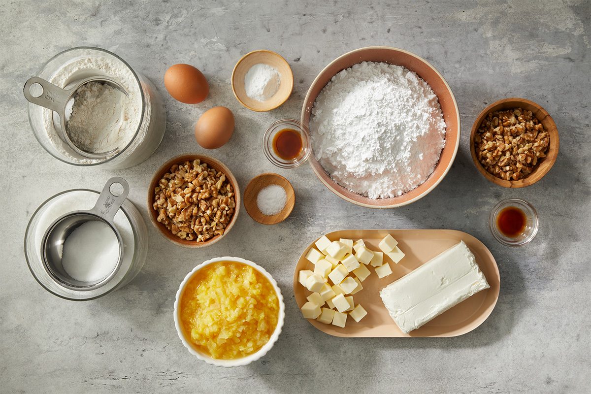 Baking ingredients arranged on a countertop, including flour, eggs, butter, cream cheese, sugar, chopped nuts, vanilla extract, salt, baking powder, and a bowl of yellow mixture, each in separate bowls or containers.