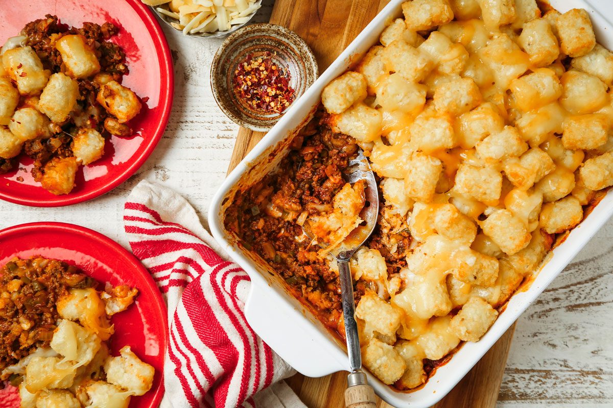 Overhead shot of Pizza Tater Tot Casserole; in a baking dish over a wooden board; served on red plates; a serving spoon and a napkin are nearby; all set on a wooden surface;
