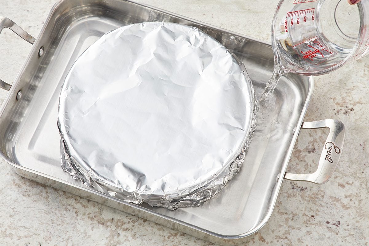 A foil-covered round pan sits inside a larger roasting pan as water is being poured from a glass measuring cup into the outer pan, preparing a water bath for baking.