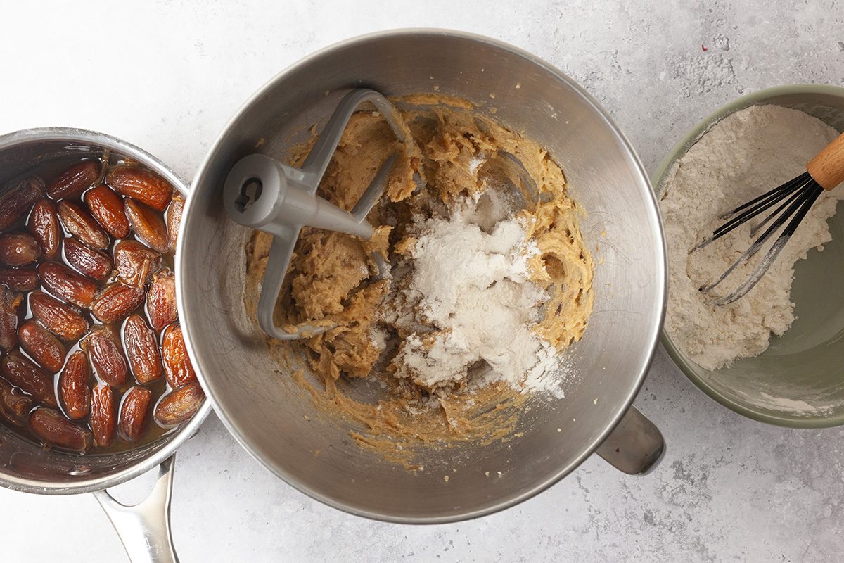 Three bowls on a countertop: one with soaked dates in liquid, one with partially mixed cookie dough and flour in a mixer bowl, and one with flour being whisked.