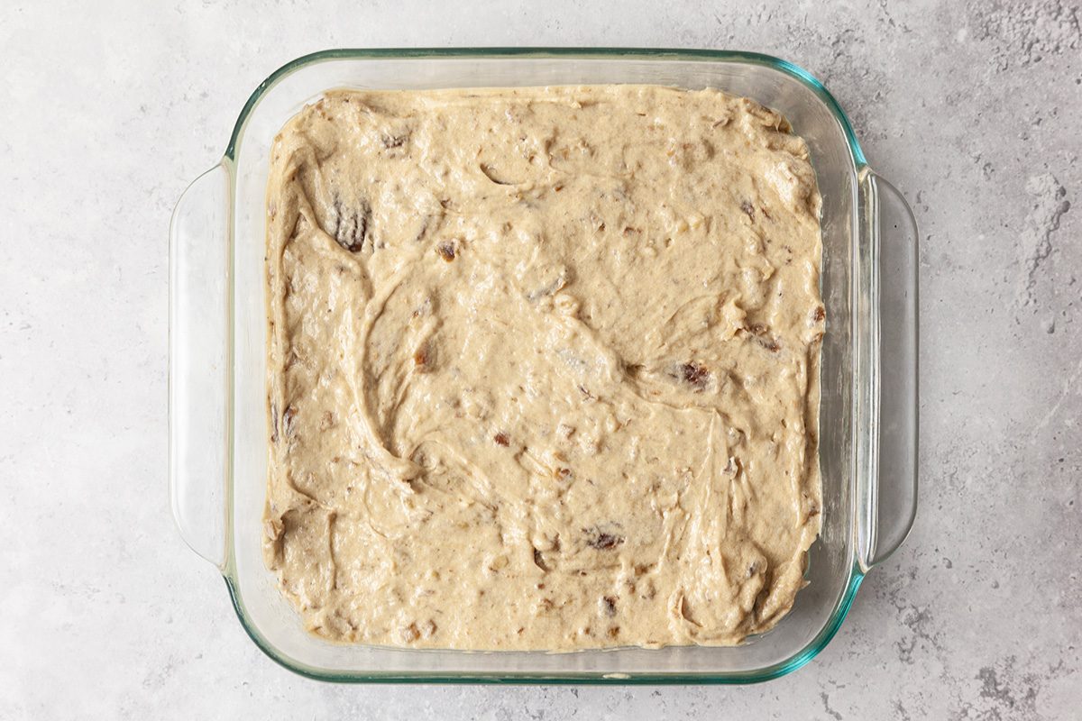 A glass baking dish filled with unbaked cookie dough, spread evenly across the bottom. The dough has a creamy texture with visible chocolate chunks and sits on a light gray countertop.