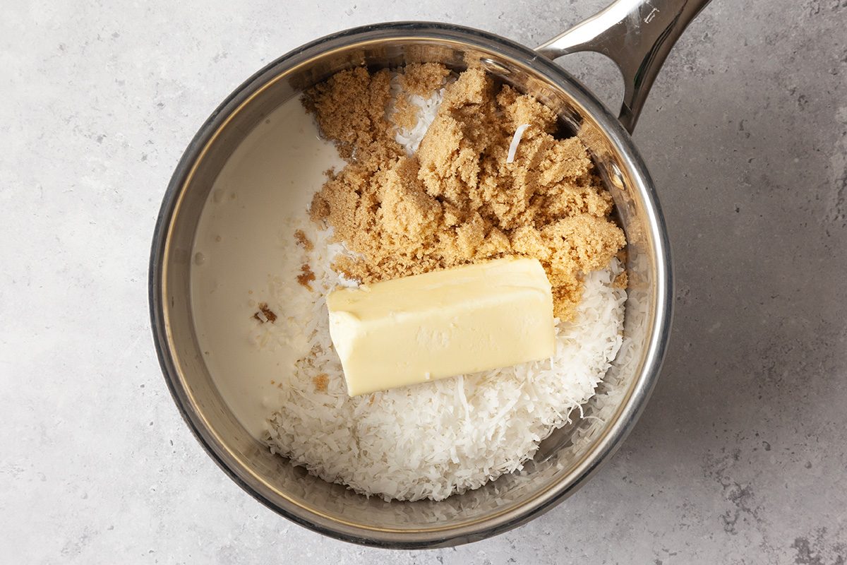A saucepan containing shredded coconut, brown sugar, and a stick of butter, all unmixed, on a light grey countertop.
