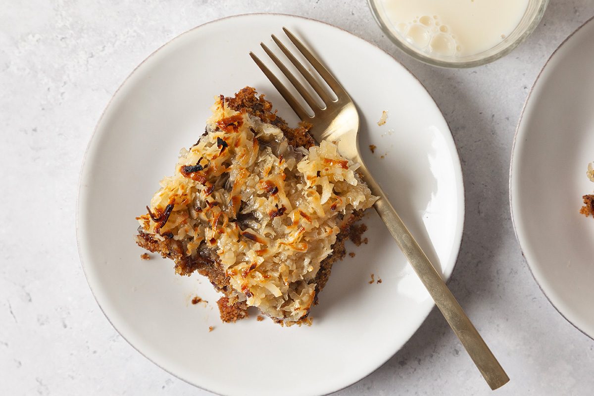 A slice of dessert with a crumbly golden-brown topping sits on a white plate with a fork, next to a glass of milk on a light surface.