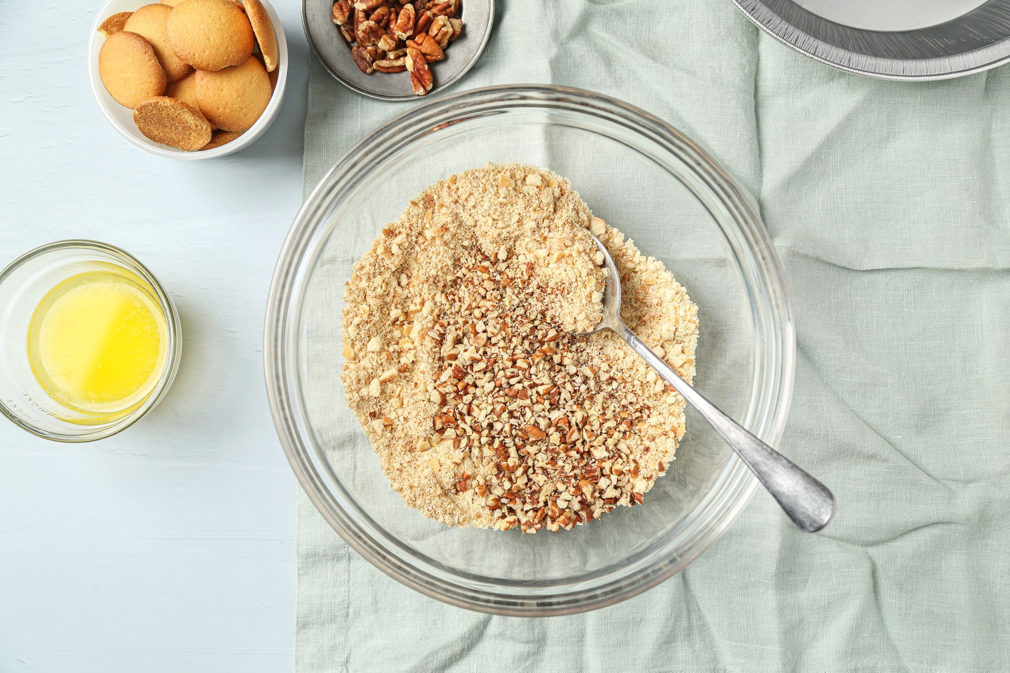 overhead shot of a glass bowl with crushed cookies and chopped nuts being mixed with a spoon, next to a bowl of vanilla wafers, a dish of pecans, melted butter, and an empty pie pan on a light blue cloth