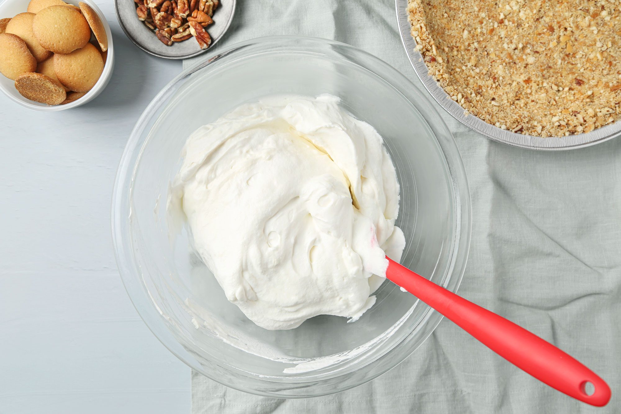 overhead shot of a glass bowl filled with whipped cream cheese mixture and a red spatula sits on a gray cloth next to bowls of vanilla wafers, chopped pecans, and a prepared graham cracker crust