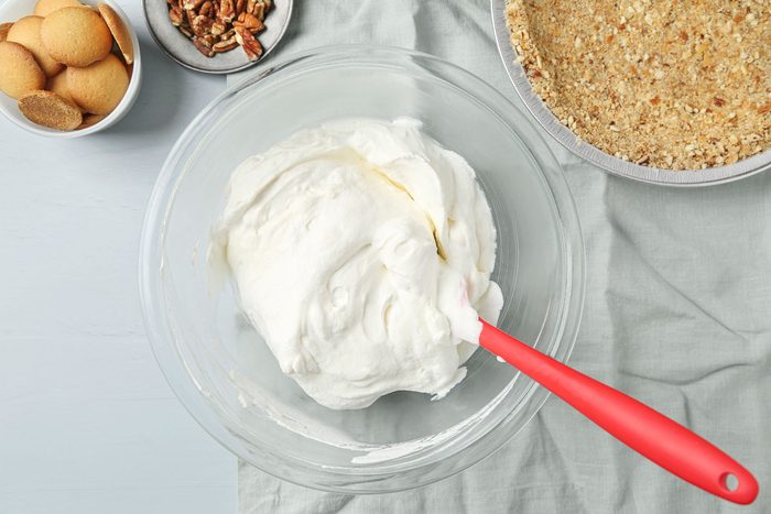 overhead shot of a glass bowl filled with whipped cream cheese mixture and a red spatula sits on a gray cloth next to bowls of vanilla wafers, chopped pecans, and a prepared graham cracker crust