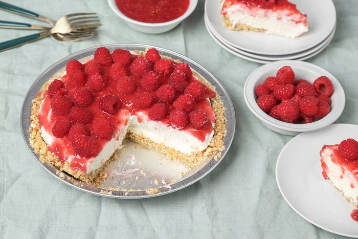 3/4th shot of a cheesecake topped with fresh raspberries and glaze sits on a table, with a slice missing, Plates with cheesecake slices, bowls of raspberries, and a bowl of raspberry sauce are also visible