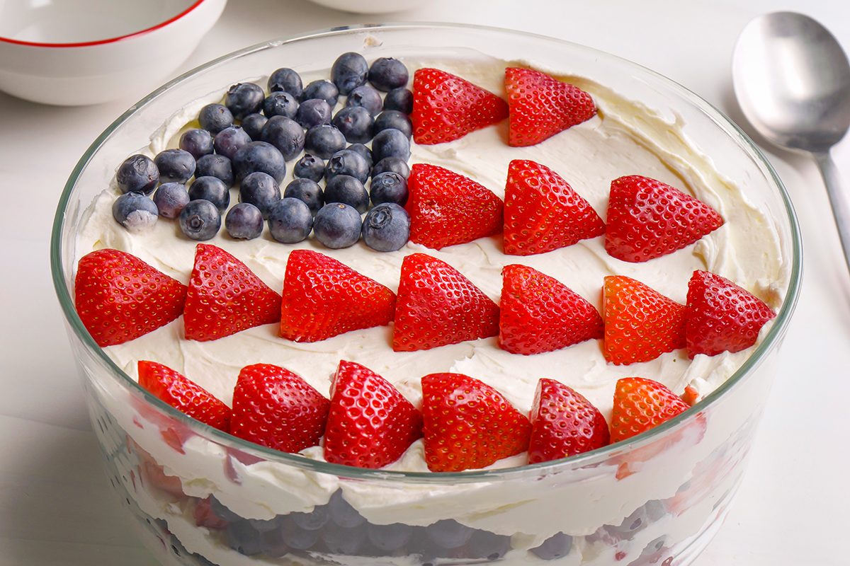 A dessert trifle topped with whipped cream, blueberries arranged in a square, and rows of halved strawberries, resembling the American flag. A spoon and bowls are visible in the background.