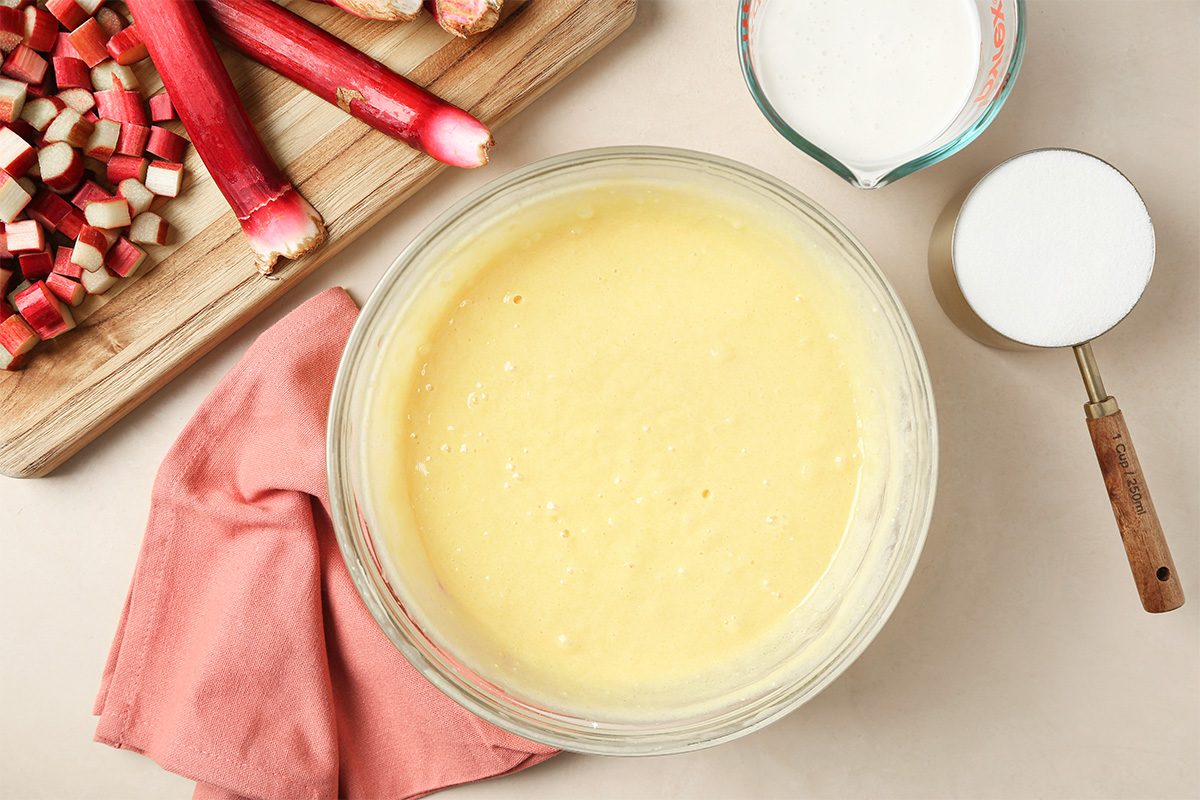 A glass bowl filled with yellow cake batter sits on a counter next to chopped rhubarb on a cutting board, a pink cloth, a cup of sugar, and a small bowl of white powder.