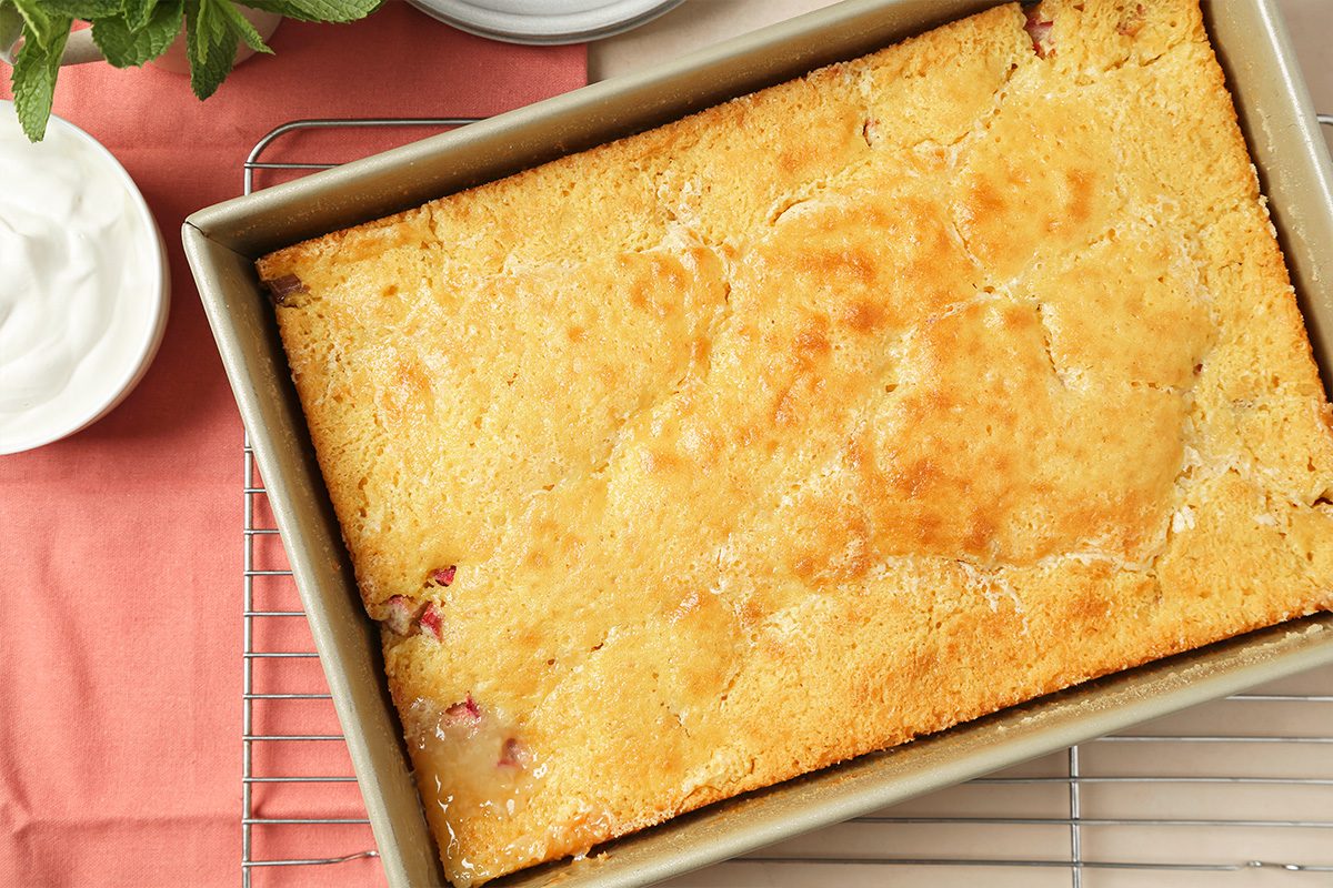 A golden-brown baked cake in a rectangular metal pan rests on a cooling rack, with a bowl of whipped cream and a pink napkin nearby.