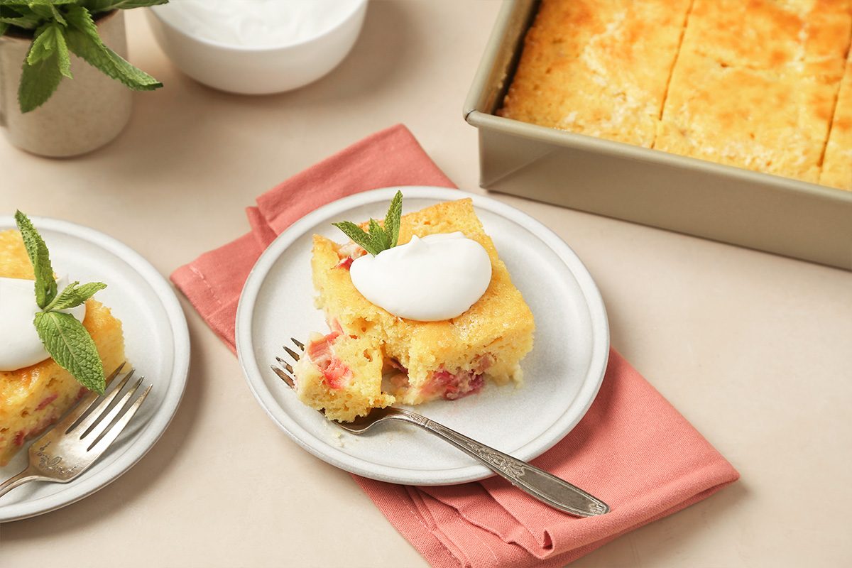 A slice of yellow cake with visible fruit pieces, topped with whipped cream and a mint sprig on a white plate with a fork, next to a baking dish of cake on a pink napkin.