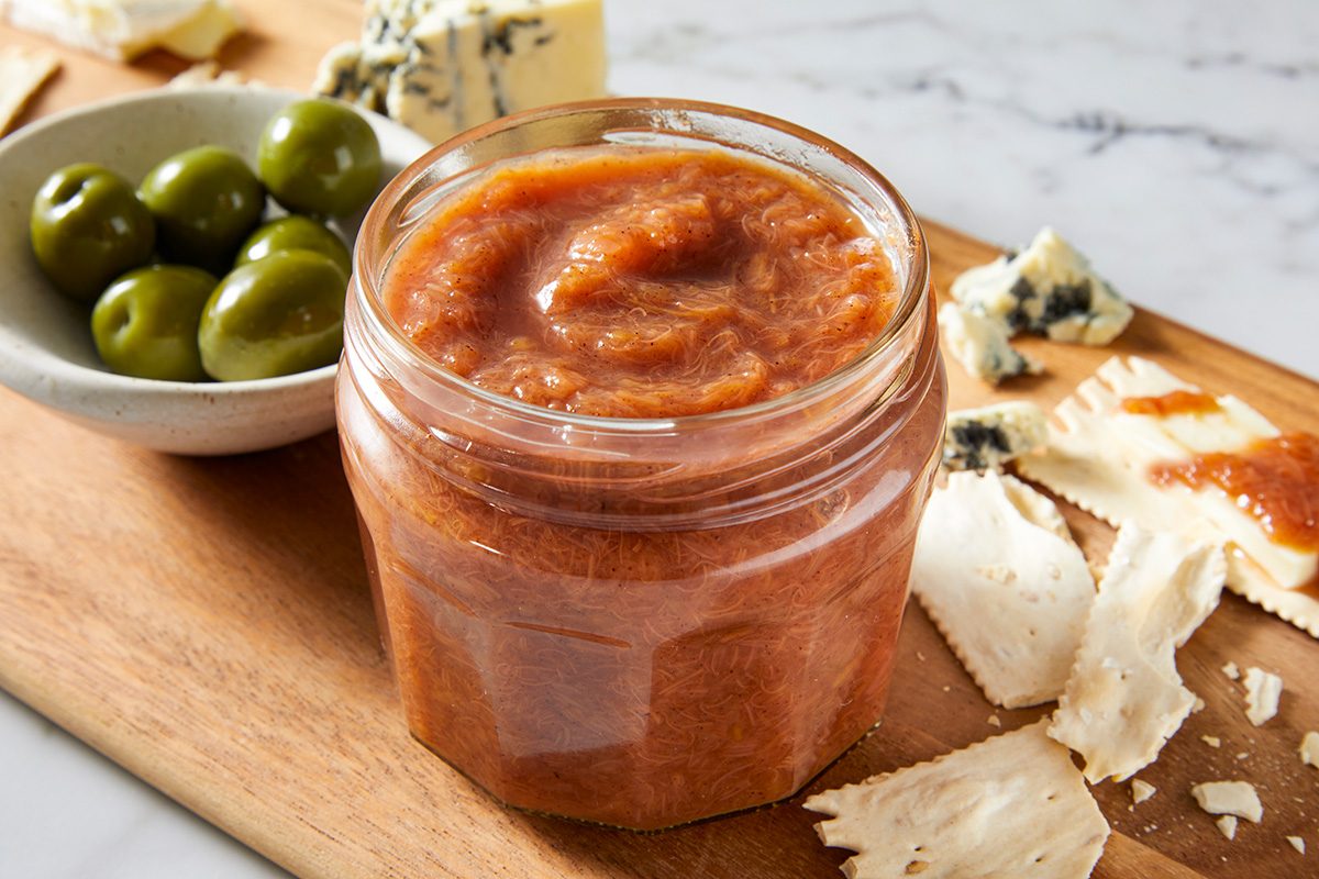 A jar of fig spread sits on a wooden board next to green olives in a bowl, crumbled blue cheese, and pieces of cracker on a marble surface.