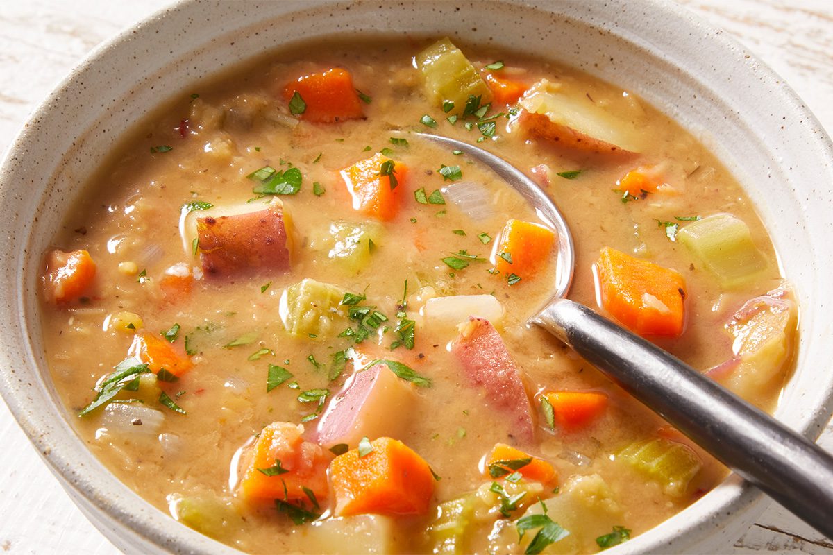 A bowl of hearty vegetable soup with chunks of carrots, potatoes, celery, and herbs, with a spoon resting inside the bowl.