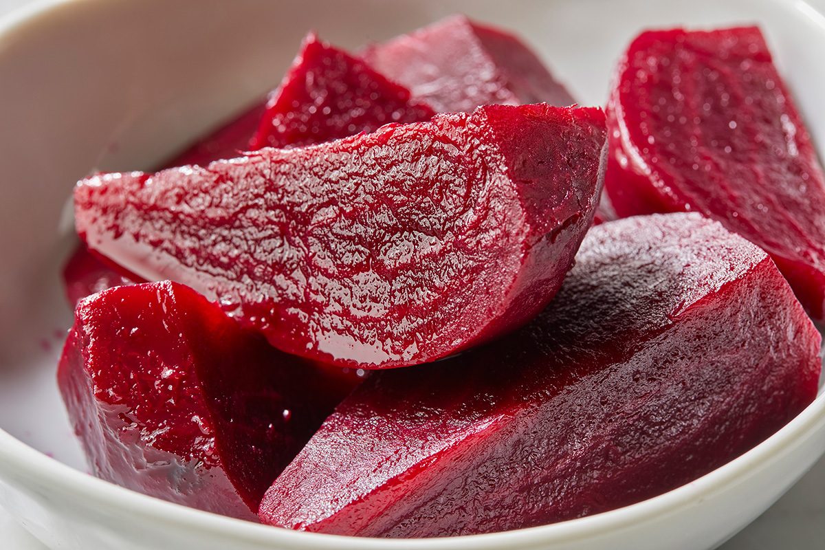 Close-up of several bright red, cooked beetroot wedges arranged in a white bowl. The beets look glossy and moist, highlighting their smooth texture and rich color.
